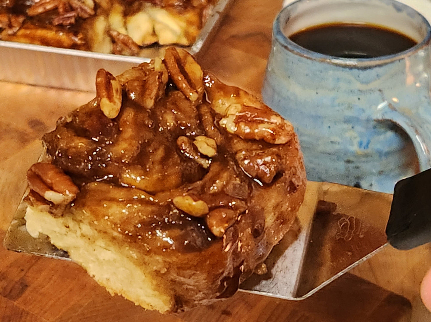 A close-up of a sticky caramel pecan roll on a metal spatula, with gooey caramel and whole pecans on top. In the background, there's a blue ceramic mug filled with black coffee and a tray of more caramel rolls on a wooden surface.