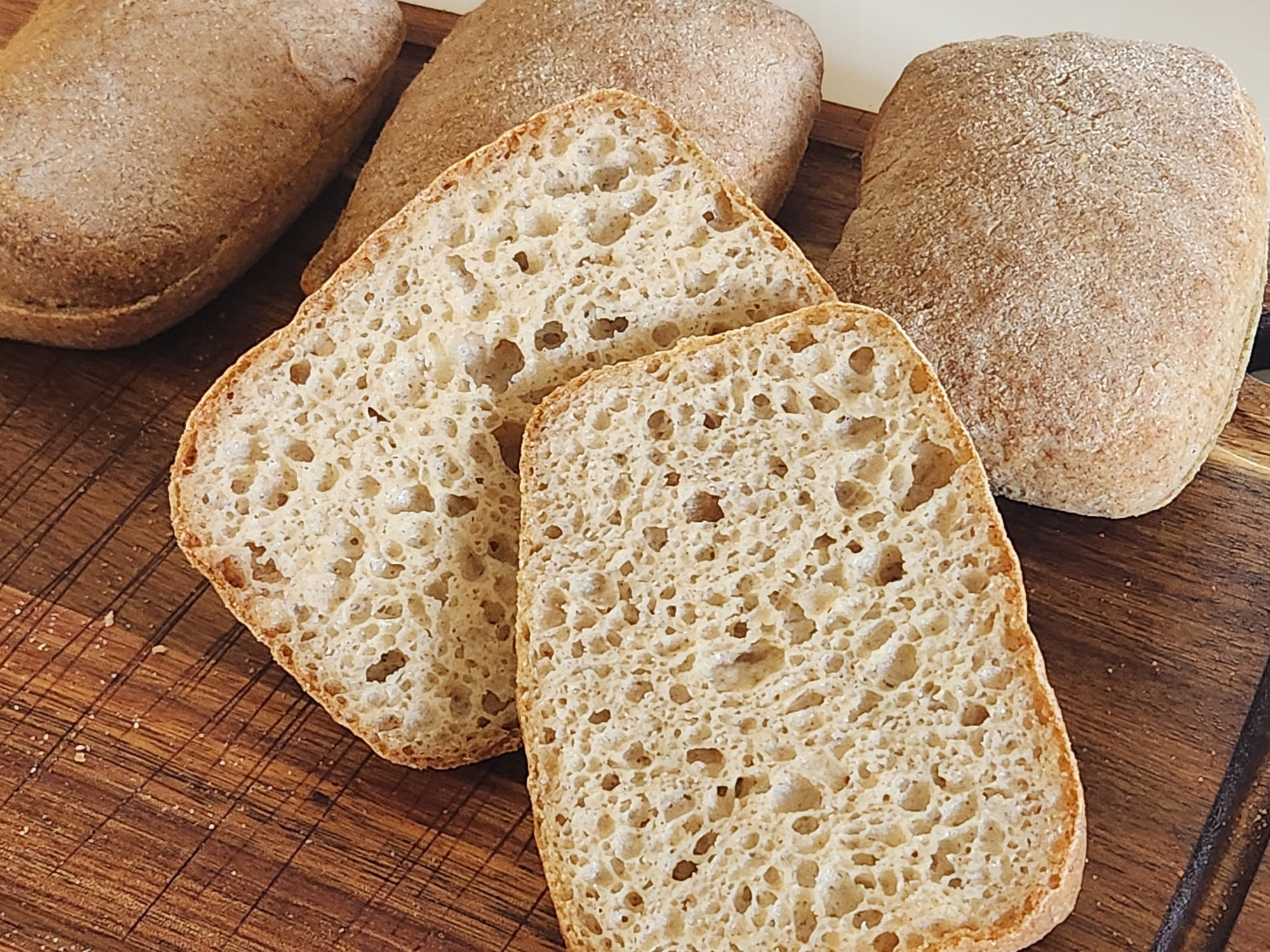 A close-up photograph of freshly baked ciabatta rolls on a wooden cutting board. One of the rolls is sliced in half, revealing its airy, open crumb structure with an even distribution of small and large holes. The crust is golden-brown with a slightly rough texture, while the interior appears soft and light. The background is slightly blurred, focusing attention on the bread's details.