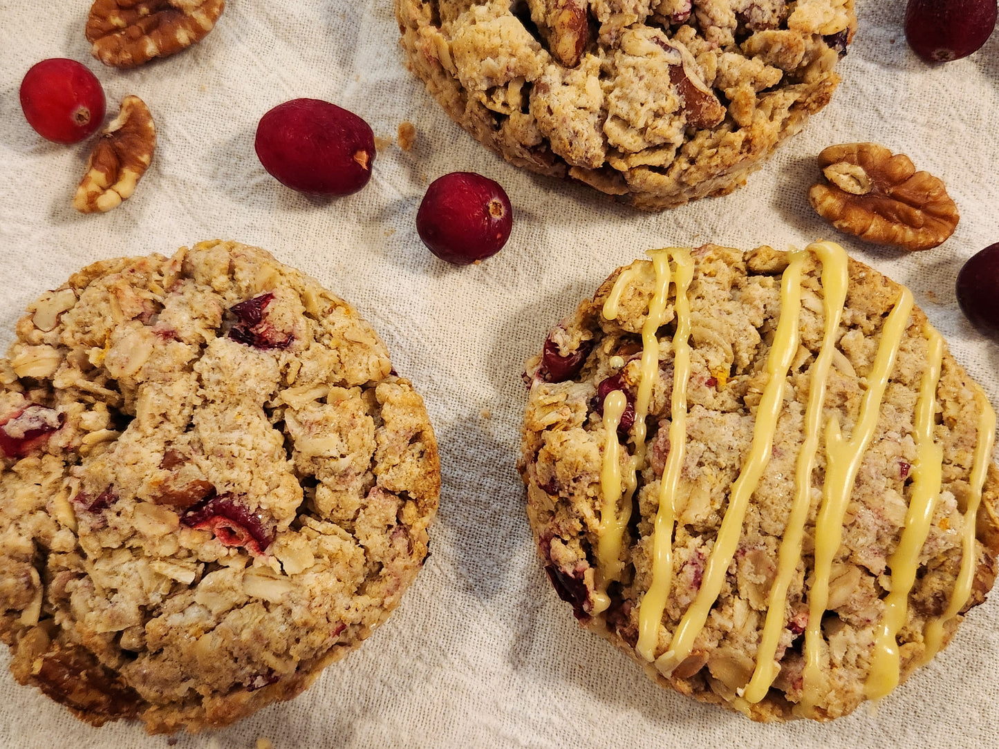 Three gluten-free cranberry orange pecan scones on a light cloth surface, surrounded by whole cranberries and pecan halves. One scone is topped with a drizzle of orange glaze, while the others display a rustic, crumbly texture with visible chunks of cranberry and pecan.