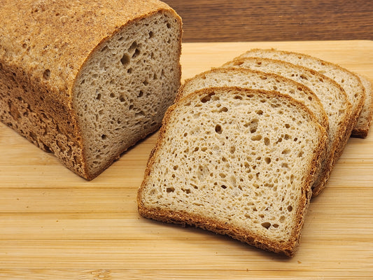 A close-up photograph of a freshly baked loaf of whole-grain bread on a wooden cutting board, with several slices cut and fanned out. The bread has a golden-brown, crusty exterior and a soft, airy interior with an open crumb structure. The texture of the crust appears slightly rough, while the slices reveal a porous and well-baked consistency. The background features a wooden surface, complementing the natural, rustic presentation of the bread.