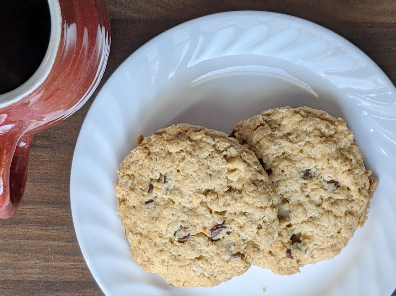 Top-down view of two oatcakes on a white plate, placed on a wooden surface next to a red ceramic mug filled with coffee. The un-iced oatcakes have a rough, hearty texture with visible bits of nuts and oats."