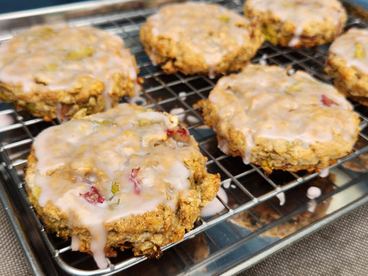 Iced oatmeal scones with visible bits of red rhubarb are arranged on a wire cooling rack over a metal tray. The scones are coated with a thin, glossy glaze that drips slightly down the sides.