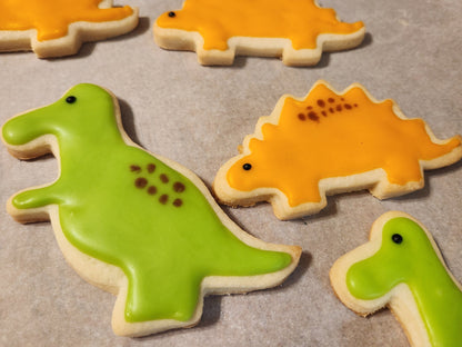 A set of decorated gluten-free sugar cookies shaped like various dinosaurs, including a triceratops and stegosaurus with green and orange icing.