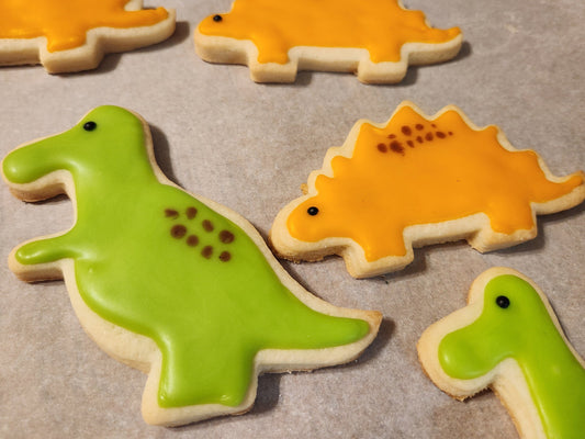 A set of decorated gluten-free sugar cookies shaped like various dinosaurs, including a triceratops and stegosaurus with green and orange icing.