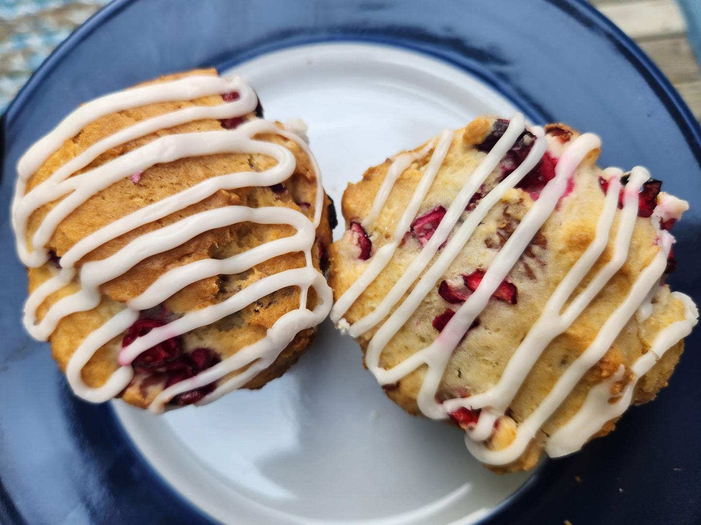 Overhead view of two muffins studded with red cranberries are shown on a white plate with a dark blue rim. The scones are generously drizzled with white icing in zigzag patterns across the top.