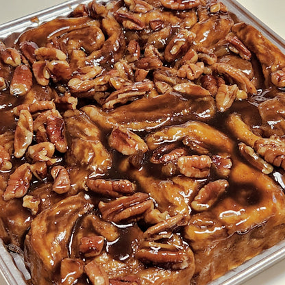 A close-up view of a tray of sticky caramel pecan rolls. The rolls are generously topped with glossy caramel sauce and a large amount of chopped and whole pecans, creating a rich, indulgent appearance.