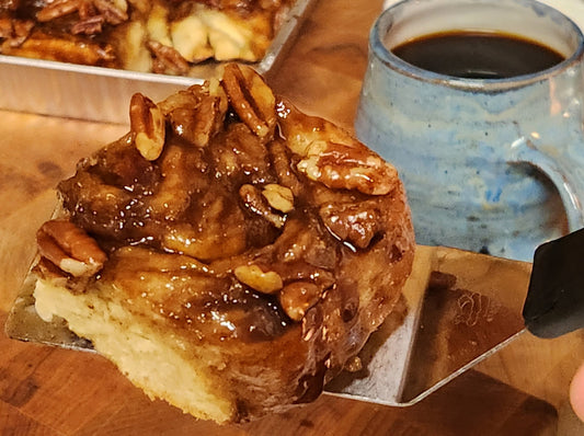 A close-up of a sticky caramel pecan roll on a metal spatula, with gooey caramel and whole pecans on top. In the background, there's a blue ceramic mug filled with black coffee and a tray of more caramel rolls on a wooden surface.
