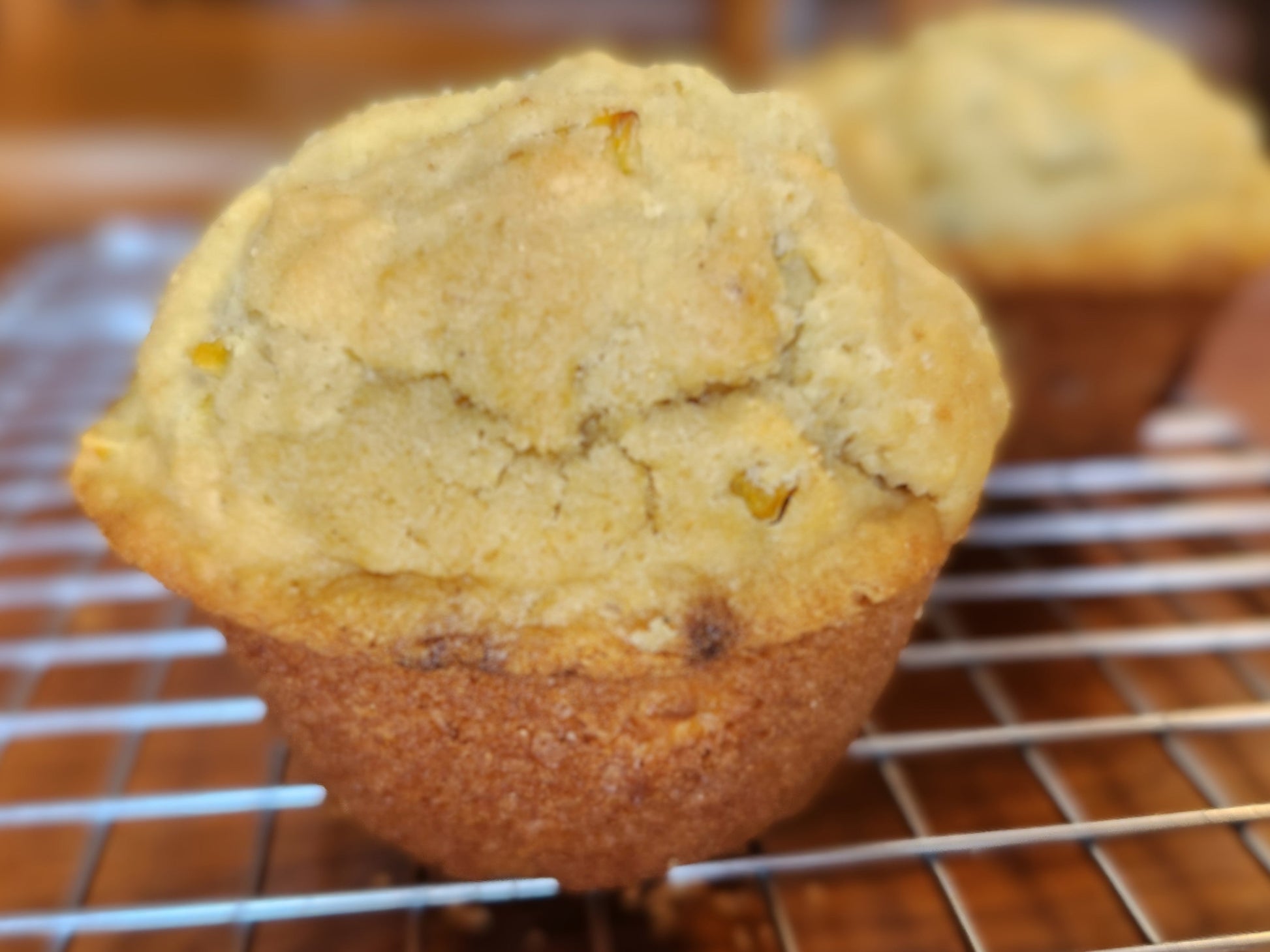 A close-up photograph of a freshly baked cornbread muffin with a golden-brown crust, resting on a wire cooling rack. The muffin has a slightly domed top with a cracked surface, revealing bits of yellow corn kernels inside. In the background, additional muffins are slightly out of focus. The setting appears to be a kitchen with warm lighting.