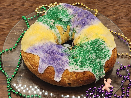 This image features a King Cake decorated in traditional Mardi Gras colors—green, gold, and purple. The cake is iced and sprinkled with colored sugar, and there are Mardi Gras beads and a small plastic baby figurine beside it. This is a gluten-free version of the classic King Cake, a popular treat during the Mardi Gras season.