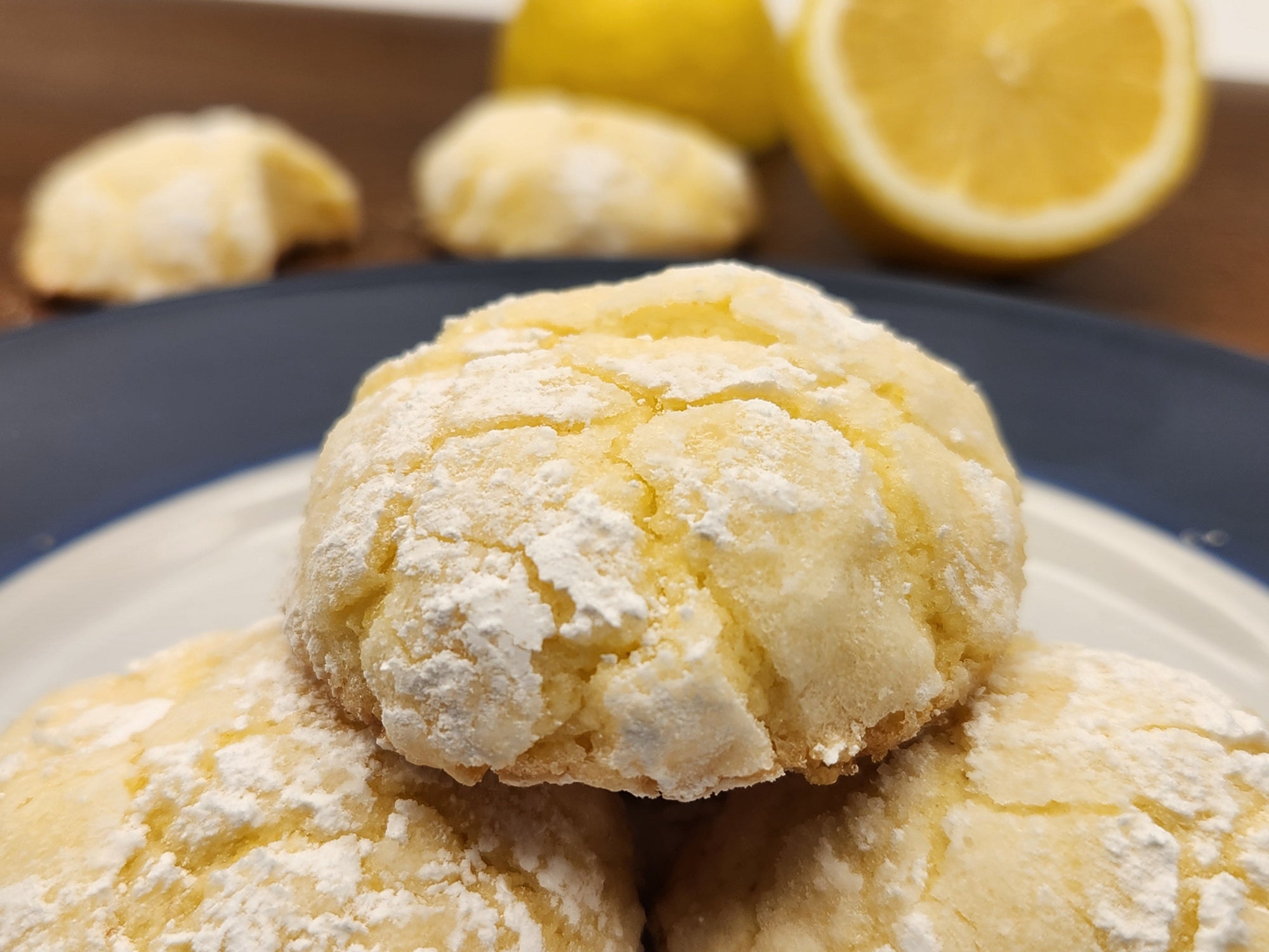 This image shows delicious lemon crinkle cookies, dusted with powdered sugar, with a soft, cracked surface. The background features fresh lemons, reinforcing their citrusy flavor.
