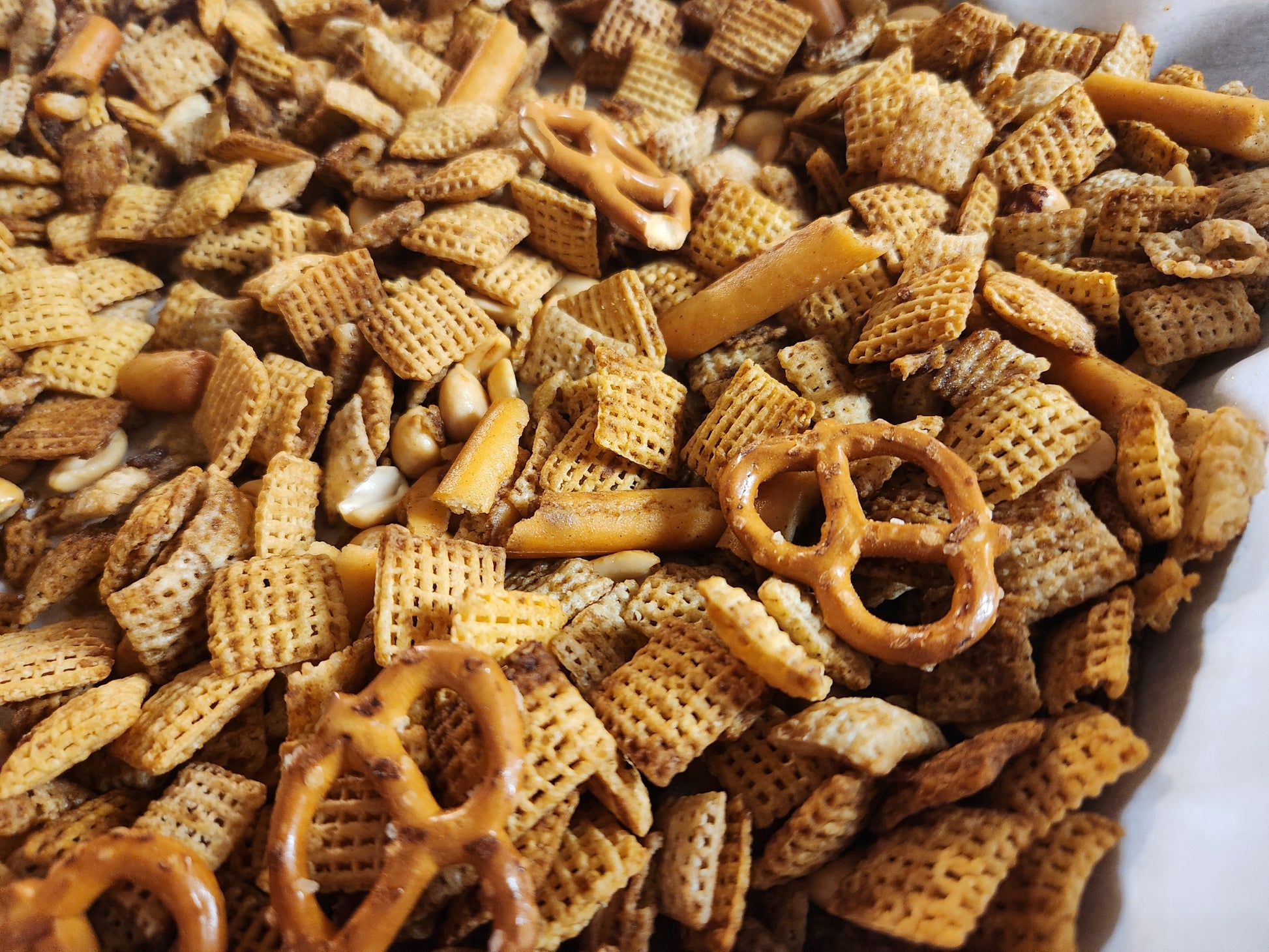 A close-up photograph of a homemade gluten-free party mix featuring crispy rice and corn square-shaped cereal, small pretzels, peanuts, and crunchy breadstick pieces. The mix is coated in a savory seasoning, giving it a golden-brown hue. The textures of the various ingredients are visible, with a balance of smooth, ridged, and knobby surfaces. The image is taken at an angle that highlights the depth and variety of the snack mix.