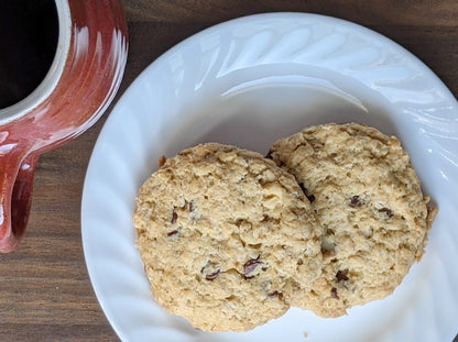 Top-down view of two oatcakes on a white plate, placed on a wooden surface next to a red ceramic mug filled with coffee. The un-iced oatcakes have a rough, hearty texture with visible bits of nuts and oats."